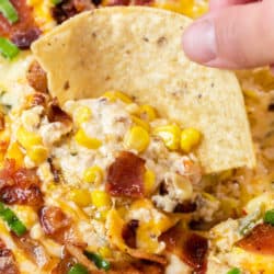 A hand holding a tortilla chip and dipping it into hot corn dip with bacon.