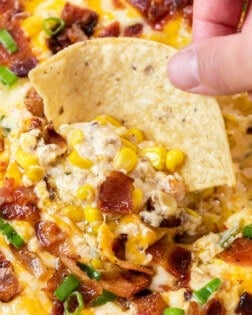 A hand holding a tortilla chip and dipping it into hot corn dip with bacon.