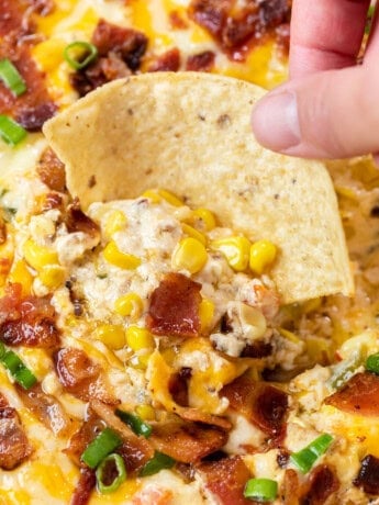 A hand holding a tortilla chip and dipping it into hot corn dip with bacon.