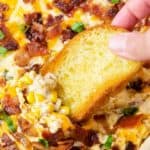 A hand dipping a baguette into cheesy corn casserole topped with bacon.