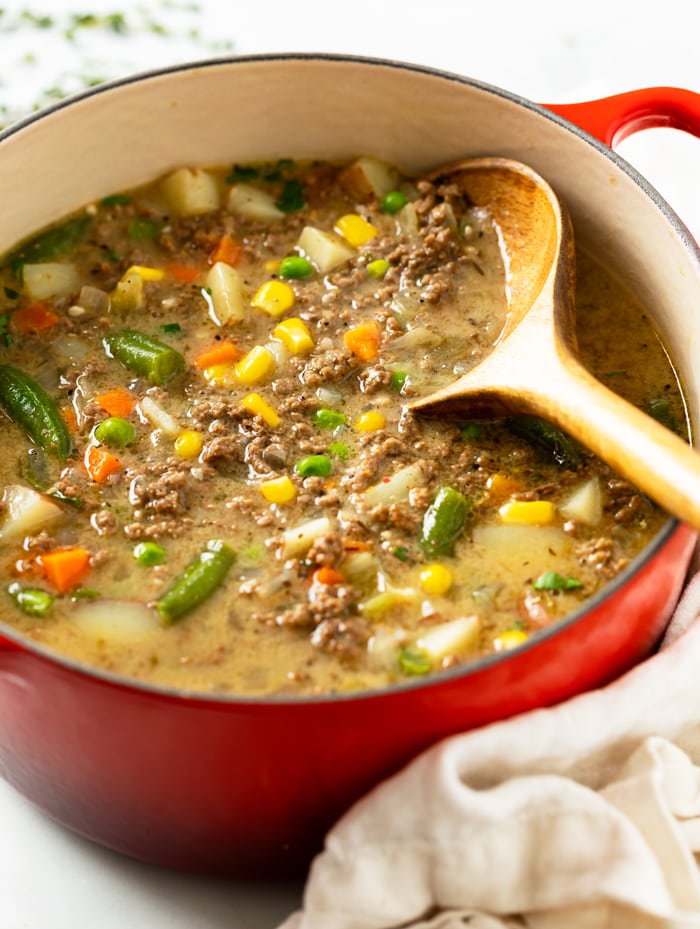 A red soup pot filled with Hamburger Soup with ground beef, vegetables, and a wooden spoon.