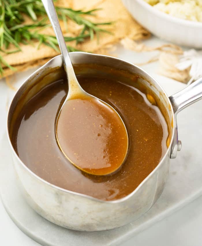 A pot of brown gravy with a spoon scooping it out with rosemary and mashed potatoes in the background.
