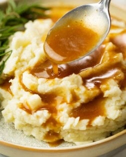 A spoon drizzling brown gravy over warm mashed potatoes.