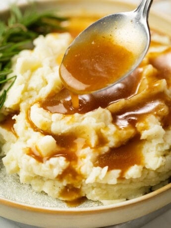 A spoon drizzling brown gravy over warm mashed potatoes.