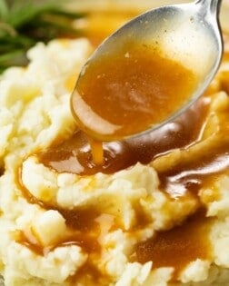 A pile of mashed potatoes on a plate with a spoon drizzling brown gravy on top.