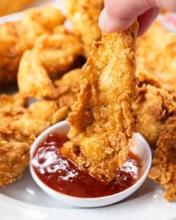 A hand holding a fried chicken tender and dipping it in sweet and sour sauce.