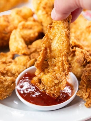 A hand holding a fried chicken tender and dipping it in sweet and sour sauce.