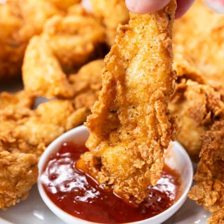 A hand holding a fried chicken tender and dipping it in sweet and sour sauce.