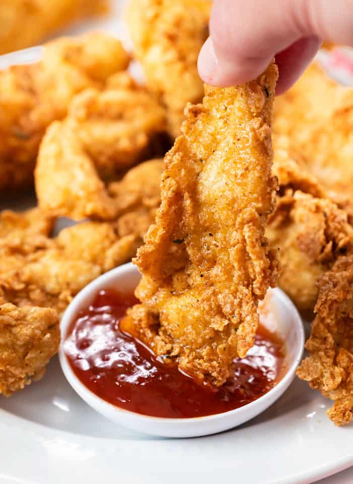 A hand holding a fried chicken tender and dipping it in sweet and sour sauce.