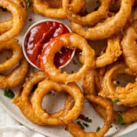 A pile of onion rings with one of them dipped into a ramekin of ketchup.