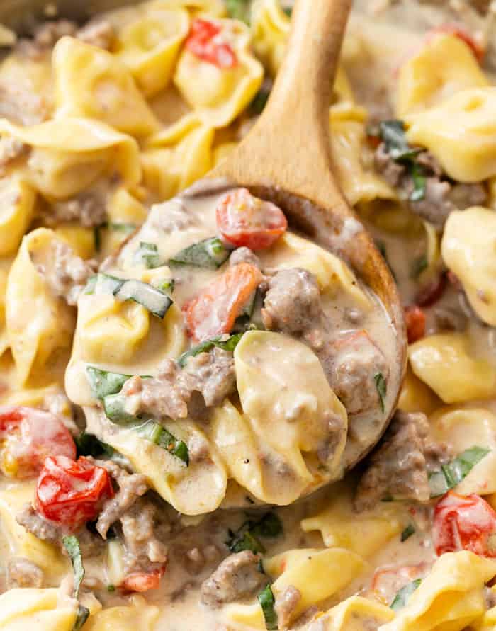 A wooden spoon scooping up creamy tomato basil tortellini pasta from a skillet.