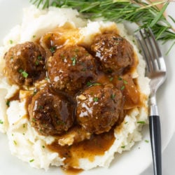 A white plate with a pile of mashed potatoes topped with meatballs and gravy with fresh rosemary on the side.