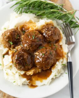 A white plate with a pile of mashed potatoes topped with meatballs and gravy with fresh rosemary on the side.