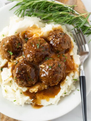 A white plate with a pile of mashed potatoes topped with meatballs and gravy with fresh rosemary on the side.