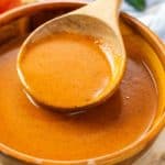 A wooden spoon scooping red enchilada sauce up from a wooden bowl.
