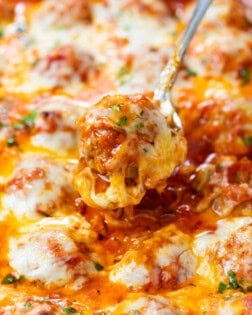 A spoon pulling up a cheesy baked meatball in marinara sauce from a casserole dish.