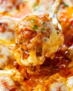 A spoon scooping up a meatball covered in marinara sauce and topped with cheese.