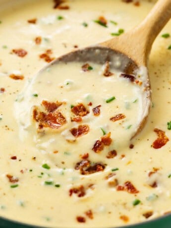 A wooden spoon scooping up baked potato soup topped with bacon and chives.