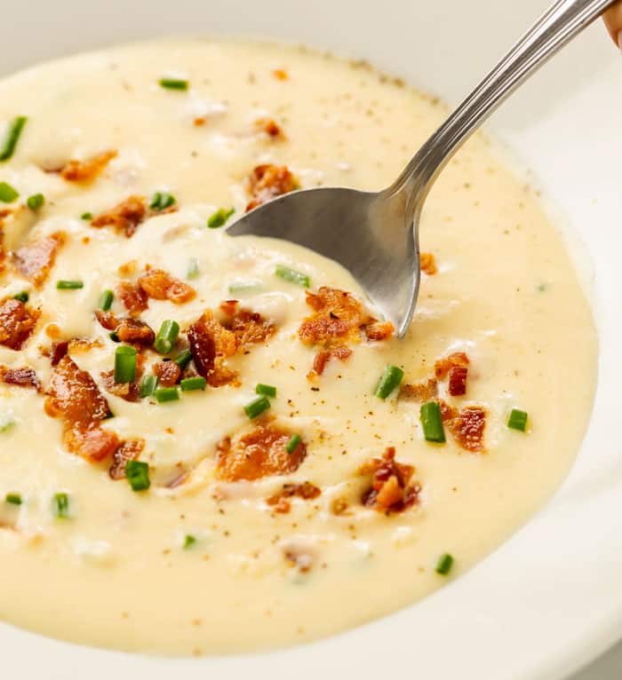 A close up view of a spoon and a white bowl with thick and creamy baked potato soup.