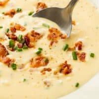 Close up view of a spoon scooping up creamy baked potato soup topped with bacon and chives.
