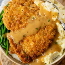 A plate with Country Fried Chicken over mashed potatoes with green beans and gravy.