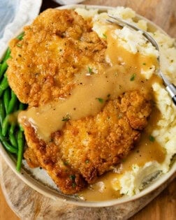 A plate with Country Fried Chicken over mashed potatoes with green beans and gravy.