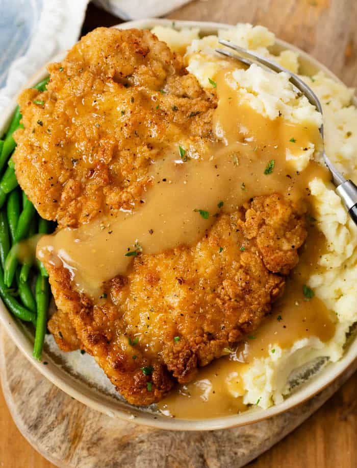 A plate with Country Fried Chicken over mashed potatoes with green beans and gravy.