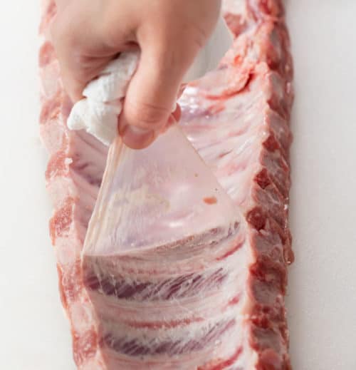 A hand pulling the membrane off of the back of baby back ribs.
