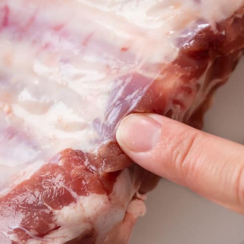 A finger pointing to a membrane on the back of a rack of baby back ribs.