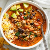 A white bowl filled with taco soup garnished with avocado and shredded cheese.