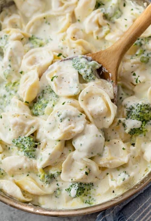 A wood spoon scooping up Tortellini Alfredo with broccoli from a skillet.