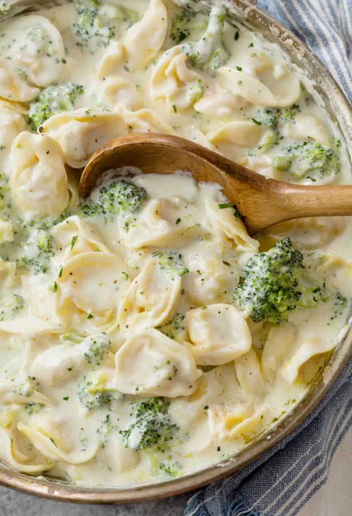 A wooden spoon in a skillet filled with Tortellini Alfredo with broccoli.