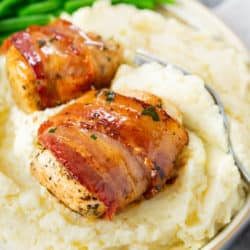 Bacon Wrapped Chicken on a pile of creamy mashed potatoes with green beans in the background.