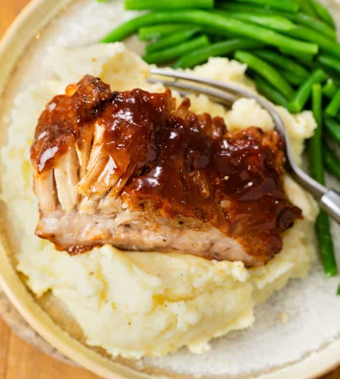 Tender Slow Cooker Ribs topped with BBQ Sauce on a pile of mashed potatoes with green beans in the background.