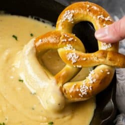 A labeled image of beer cheese dip in a cast iron skillet with a soft pretzel being dipped into it.