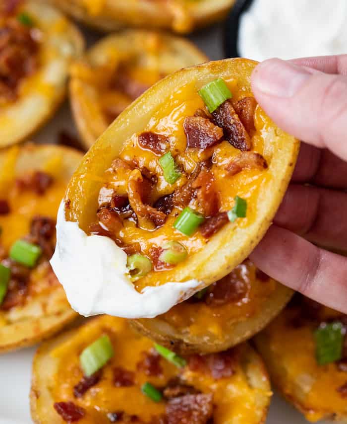 A hand holding a potato skin dipped in sour cream topped with cheese and bacon.