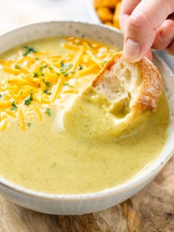 A hand dipping bread into a bowl of cream of broccoli soup.