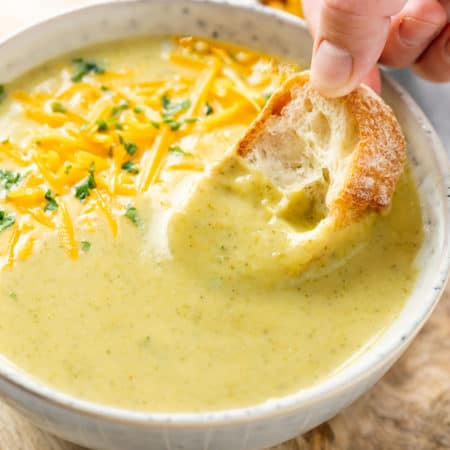 A hand dipping bread into a bowl of cream of broccoli soup.
