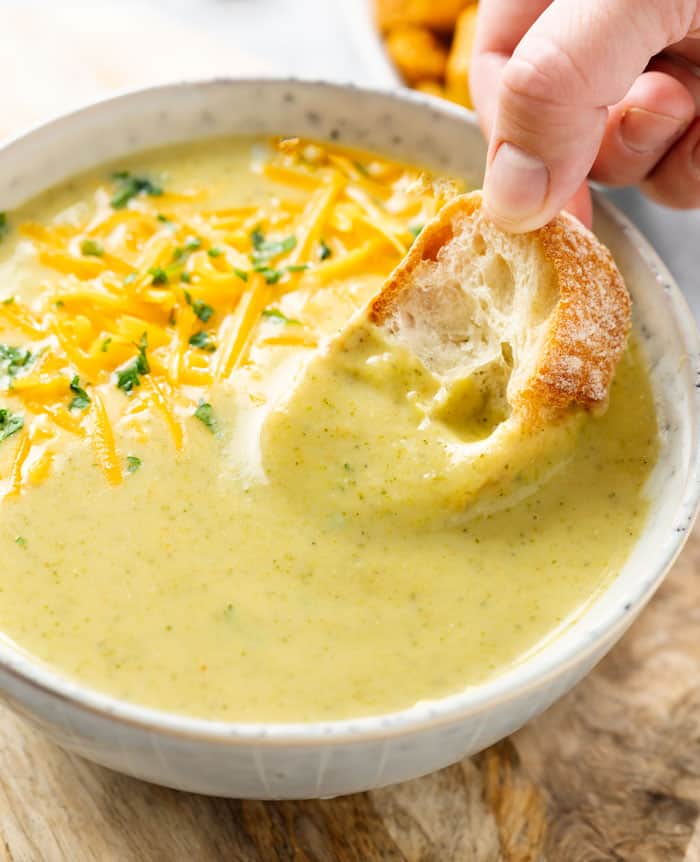 A hand dipping bread into a bowl of cream of broccoli soup.