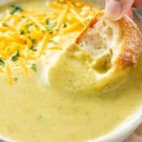A hand dipping crusty bread into a bowl of cream of broccoli soup.