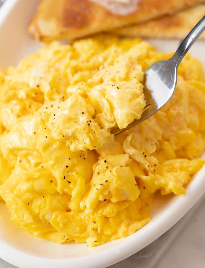 A plate of fluffy scrambled eggs with a fork scooping them up and toast in the background.