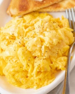 A white platter with fluffy scrambled eggs with a fork next to them and toast in the background.