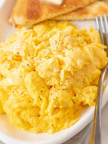 A white platter with fluffy scrambled eggs with a fork next to them and toast in the background.