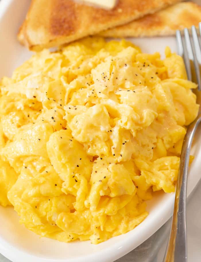 A white platter with fluffy scrambled eggs with a fork next to them and toast in the background.