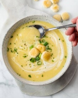 A white bowl with creamy potato leek soup in it with oyster crackers, green onions, and parsley on top.