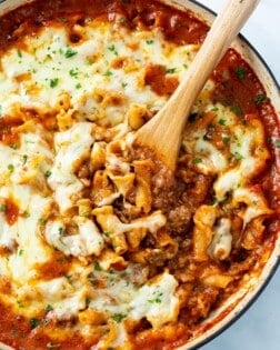 A skillet with a wooden spoon it in scooping up Skillet Lasagna topped with cheese.