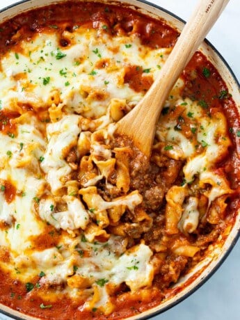 A skillet with a wooden spoon it in scooping up Skillet Lasagna topped with cheese.
