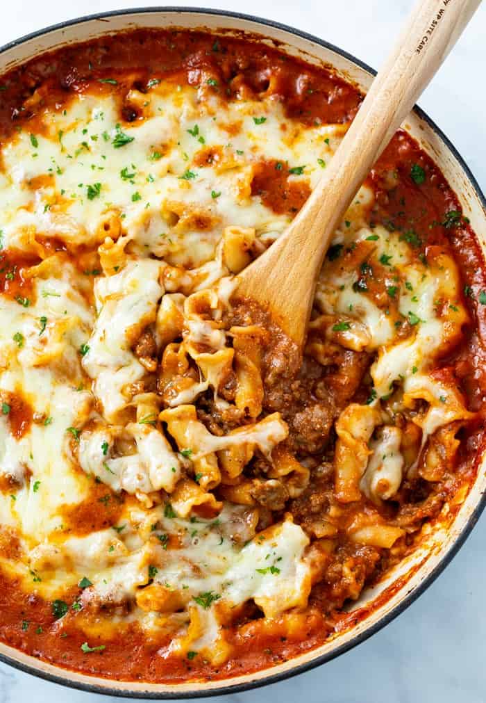 A skillet with a wooden spoon it in scooping up Skillet Lasagna topped with cheese.