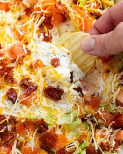 A hand dipping a potato chip into BLT Dip with Bacon, Lettuce, and Tomatoes on top.