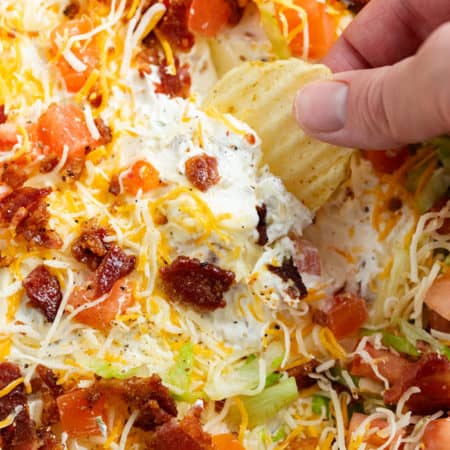 A hand dipping a potato chip into BLT Dip with Bacon, Lettuce, and Tomatoes on top.